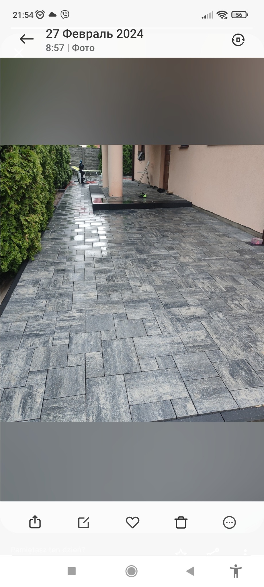 Ułożona szara kostka brukowa o nieregularnym kształcie przed domem z kolumnami, obok żywopłotu; widoczne buty i narzędzia w tle.