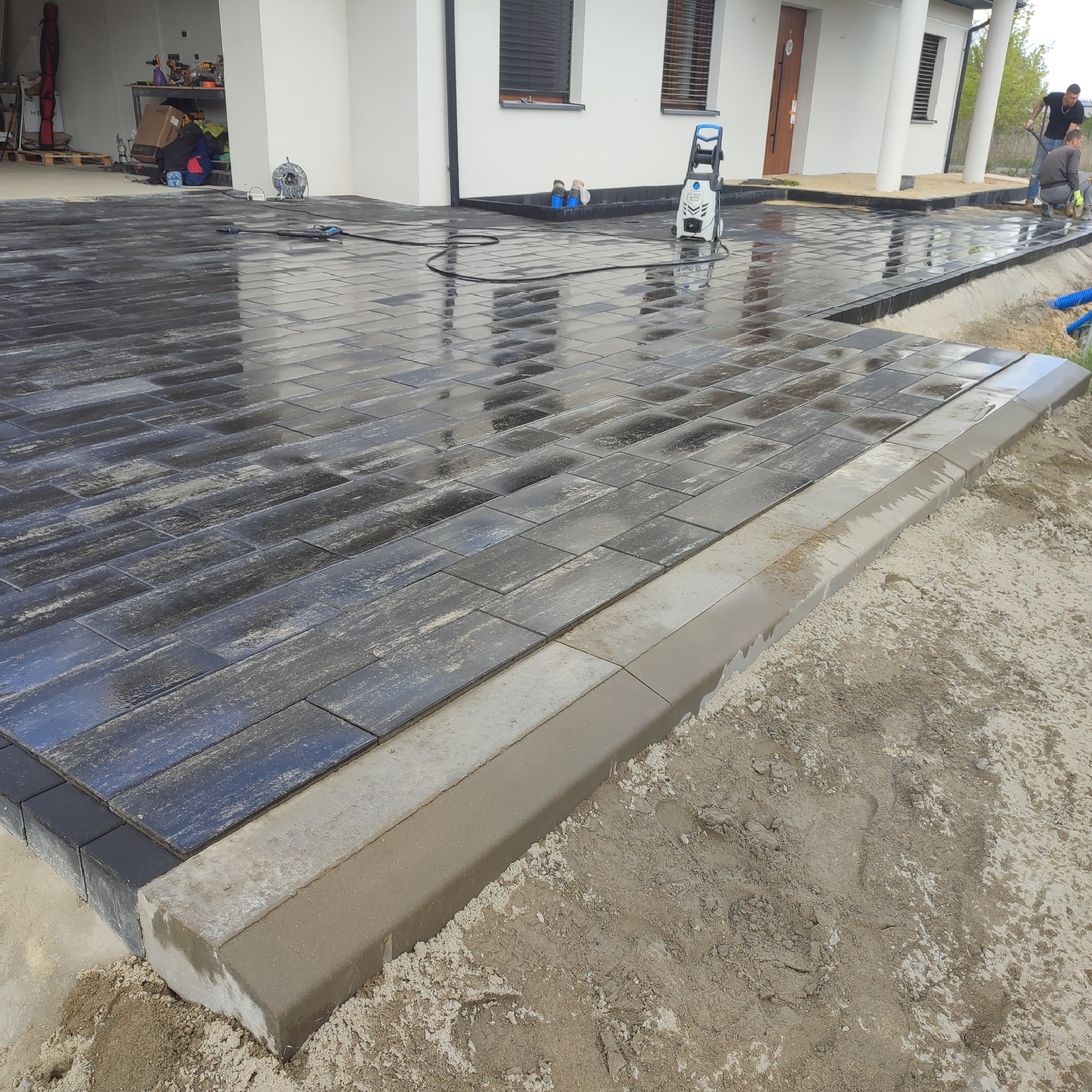 Świeżo ułożona, mokra, prostokątna kostka brukowa w odcieniach szarości, obramowana betonowym krawężnikiem. W tle biały budynek z kolumnami i pracownicy podczas czyszczenia powierzchni myjką...