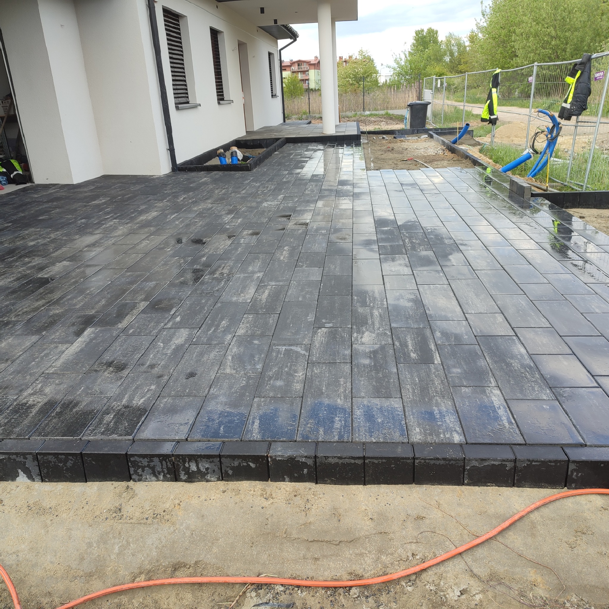 Ułożona, mokra, grafitowa kostka brukowa przed nowoczesnym domem z białymi kolumnami, widoczne narzędzia i zabezpieczenia budowlane.