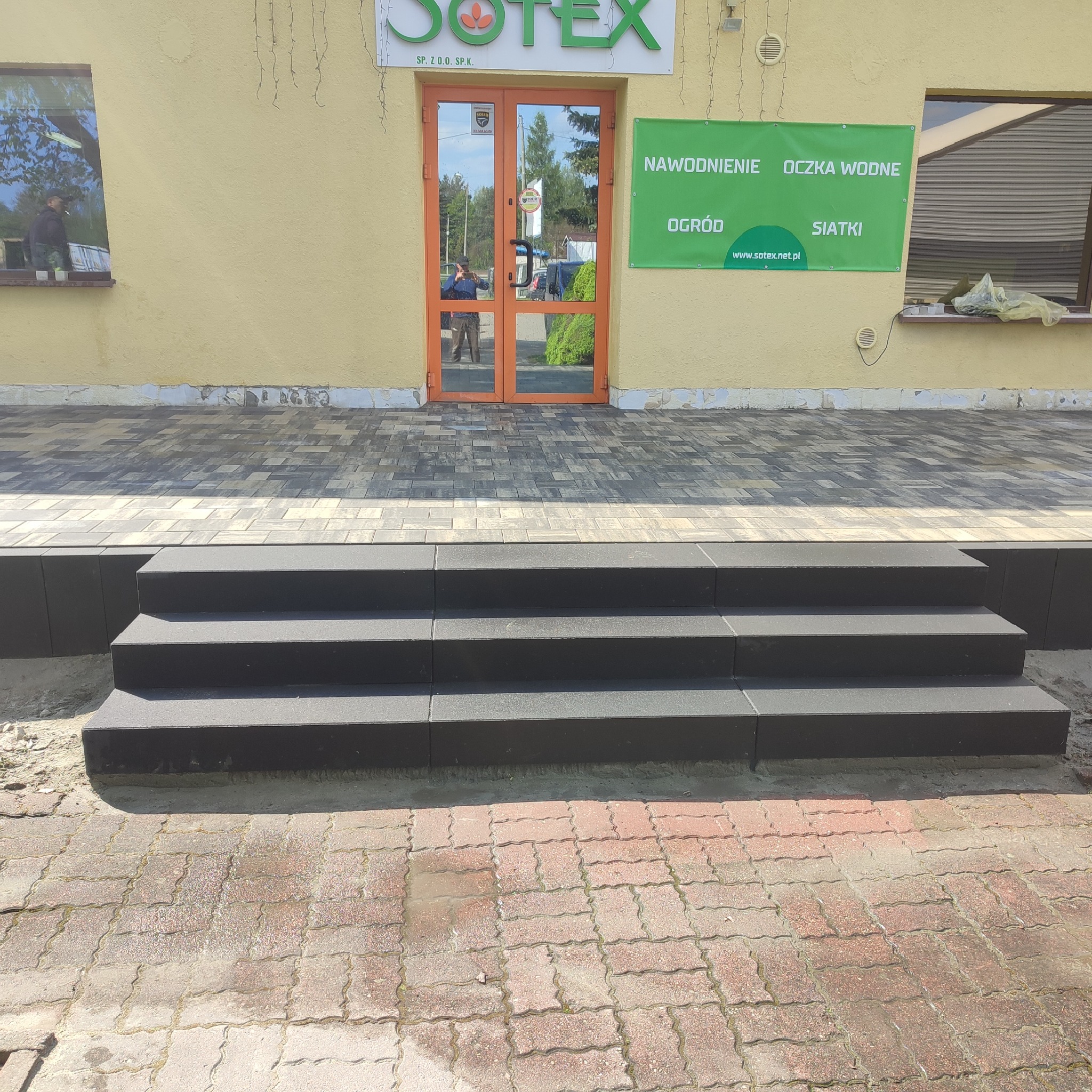 Schody z czarnej kostki prowadzące do budynku firmy Sotex, z widoczną kostką brukową w różnych odcieniach szarości i czerwieni przed schodami.
