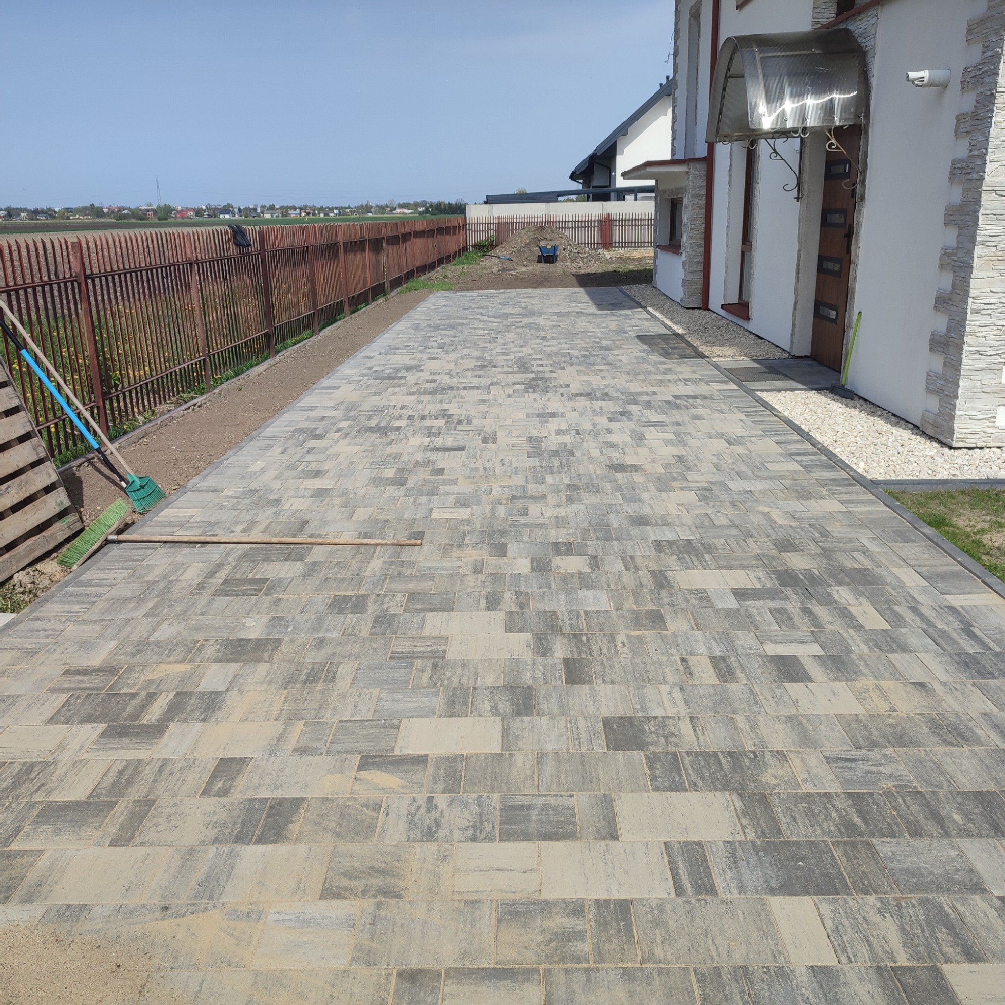 Ułożona szara kostka brukowa na podjeździe przed domem z brązowymi drzwiami, widoczny fragment ogrodzenia i narzędzia brukarskie.