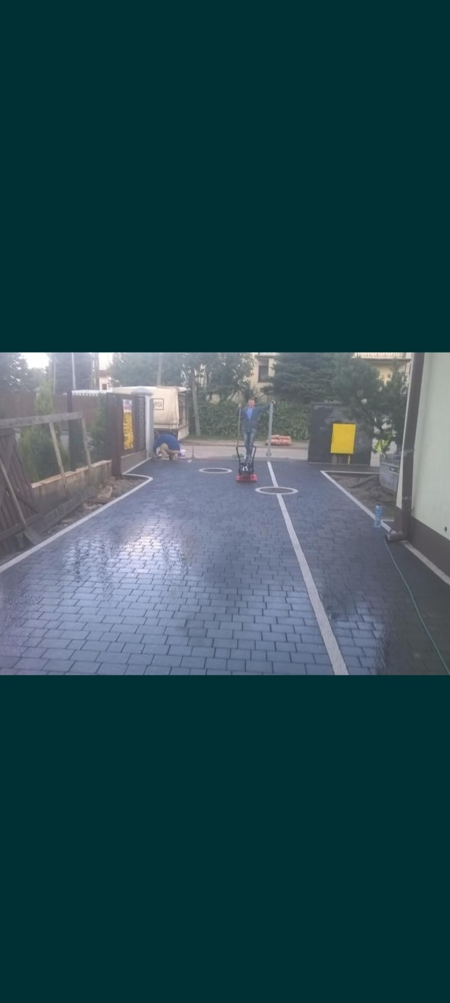 Układanie ciemnoszarej kostki brukowej na podjeździe, widoczny pracownik z zagęszczarką w trakcie pracy, obok białe linie oddzielające miejsca parkingowe.