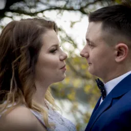Profilowe ujęcie pary młodej: panna młoda z falowanymi włosami i delikatnym makijażem oraz pan młody w granatowym garniturze i muszce w kropki, patrzący na siebie na tle drzew.