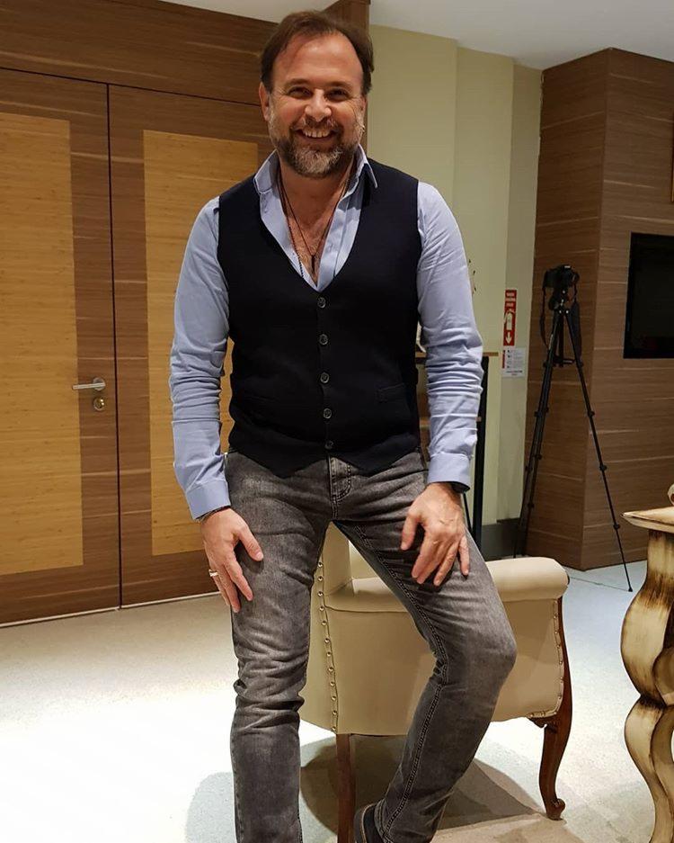 Uśmiechnięty mężczyzna w niebieskiej koszuli i czarnej kamizelce, oparty o beżowe krzesło, z kamerą na statywie w tle.