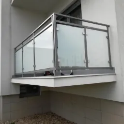 Nowoczesny balkon z matowymi szklanymi panelami i metalową ramą, widok z dołu, płytki ceramiczne na elewacji budynku.