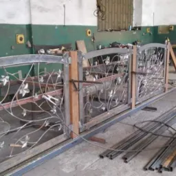 Trzy segmenty metalowej konstrukcji z motywem roślinnym, częściowo zespawane z drewnianymi słupkami, w warsztacie ślusarskim z butlą spawalniczą w tle.