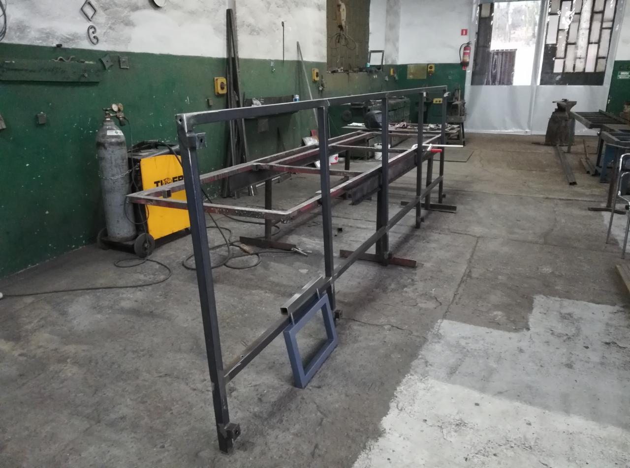 Konstrukcja metalowa w warsztacie, prawdopodobnie rama balustrady lub ogrodzenia, spawarka widoczna w tle, surowe wykończenie.