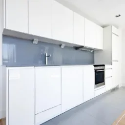 Minimalistyczna, biała kuchnia z błyszczącymi frontami szafek, szarym blatem i panelem nad blatem, widoczny fragment podłogi z szarych płytek przechodzących w drewniany parkiet.