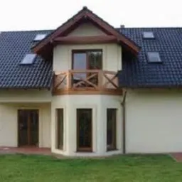 Dwukondygnacyjny dom murowany z ciemną dachówką i oknami dachowymi, z półokrągłym balkonem z drewnianą balustradą, widziany z perspektywy trawnika.