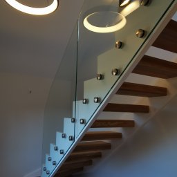 Camform Schody - Nowoczesne schody z drewnianymi stopniami i szklaną balustradą mocowaną na metalowe uchwyty. U góry widoczna okrągła lampa wisząca i kinkiet.