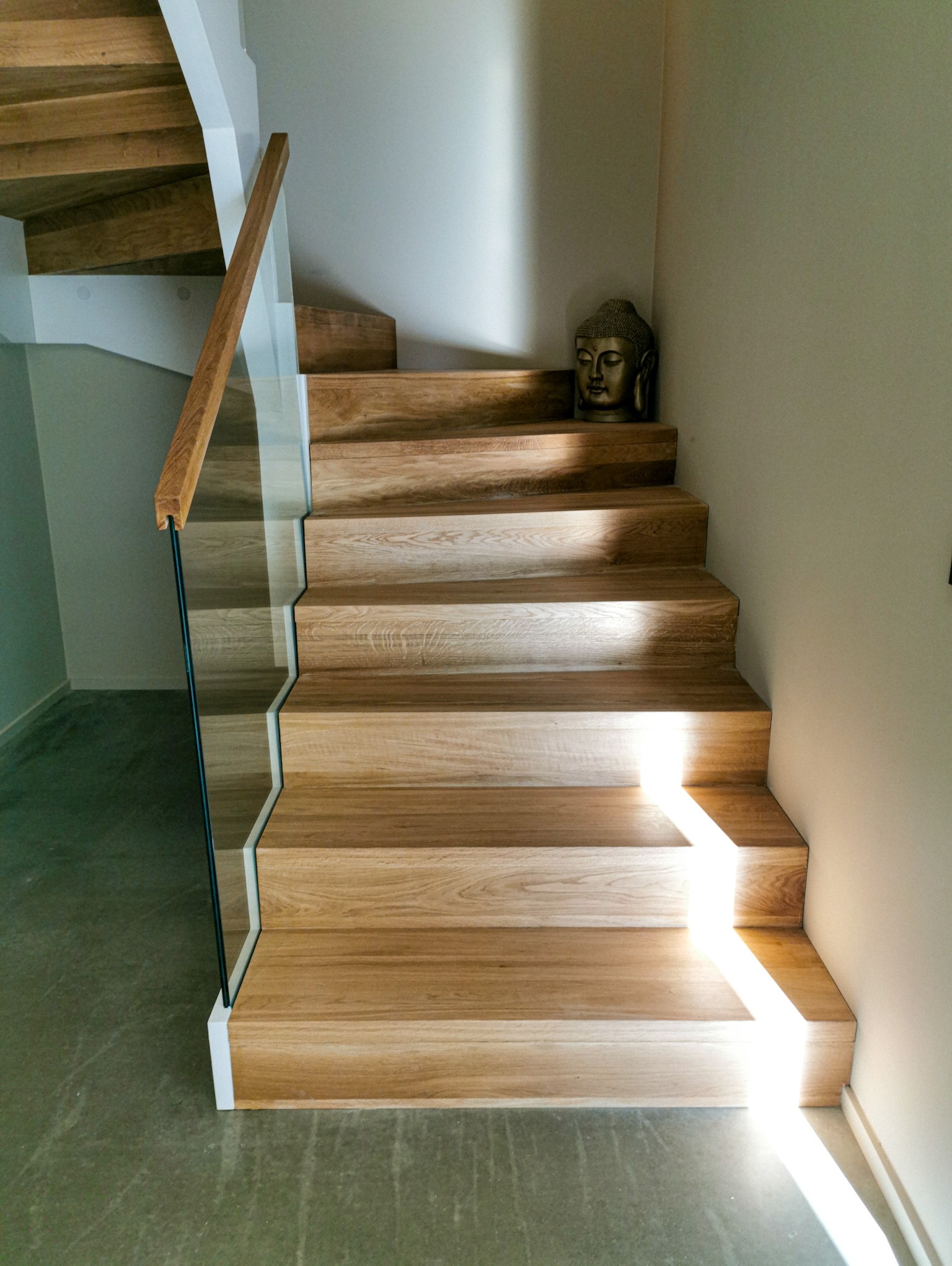 Jasne, dębowe schody z balustradą ze szkła hartowanego, bez nosków, oświetlone naturalnym światłem, z dekoracyjną figurką Buddy na jednym ze stopni.