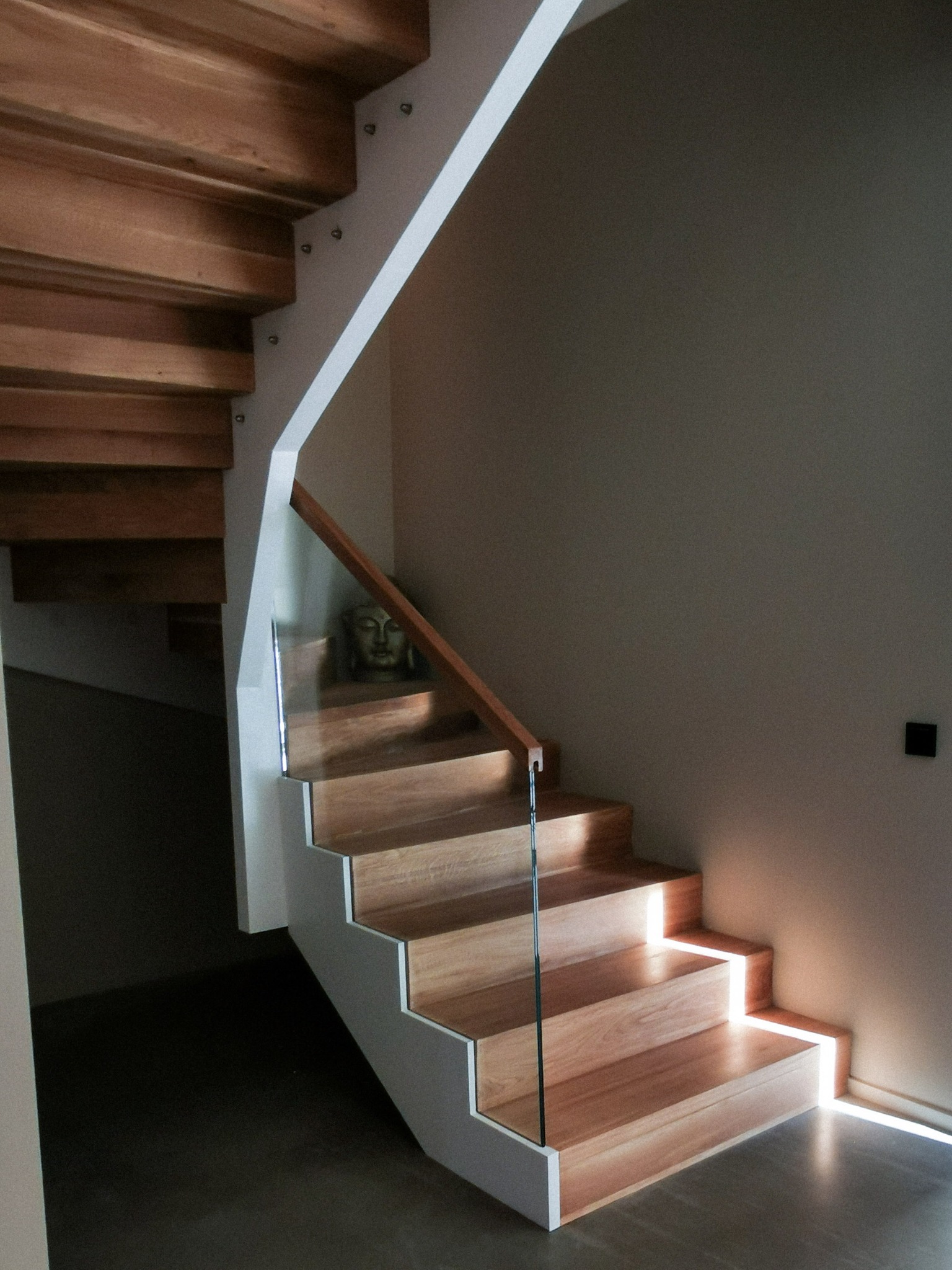 Nowoczesne schody z drewna z białymi bokami i szklaną balustradą, subtelne oświetlenie stopni, w tle widoczna statuetka Buddy.