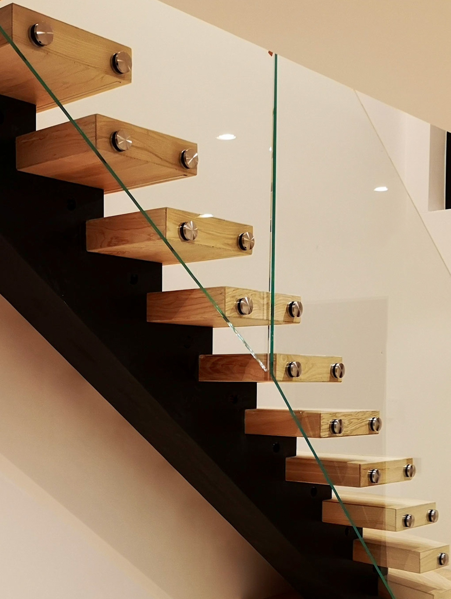 Lekkie, drewniane stopnie schodowe z dębowym fornirem, zamocowane na ciemnej, centralnej belce, zabezpieczone szklaną balustradą z metalowymi uchwytami.