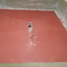 Świeżo wylana, czerwona posadzka z centralnie umieszczonym, metalowym odpływem liniowym w surowym pomieszczeniu z widocznymi śladami prac budowlanych na ścianach.