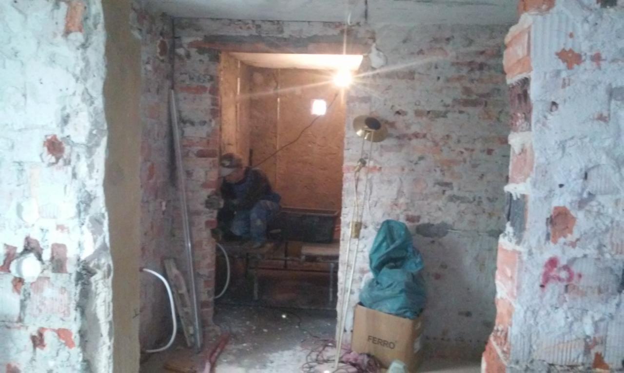 Pracownik w okularach ochronnych przygotowuje otwór drzwiowy w surowej, ceglanej ścianie. Widoczne resztki starego tynku, prowizoryczne oświetlenie halogenowe i karton Ferro.