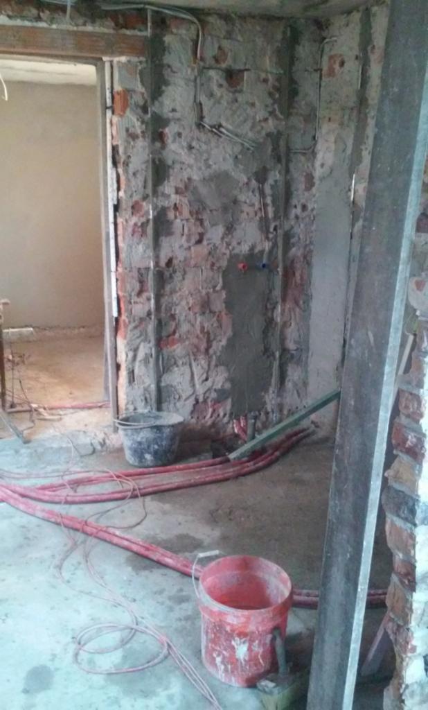 Ściana z odsłoniętą czerwoną cegłą i fragmentami szarego tynku, widoczne instalacje elektryczne i hydrauliczne, na podłodze zwinięte czerwone kable, dwa wiadra - jedno czarne, drugie czerwone,...