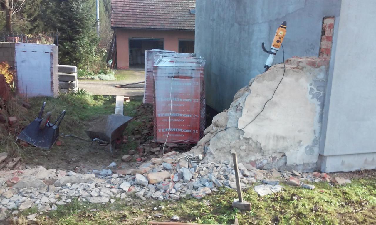 Demontaż fragmentu elewacji budynku z użyciem młota pneumatycznego, widoczne worki z bloczkami Termoton 25, gruz i dwie taczki.