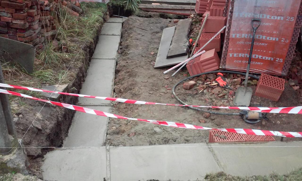 Wykop z wylanym betonem, zabezpieczony taśmą ostrzegawczą, obok sterta cegieł Termoton 25 i narzędzia budowlane.