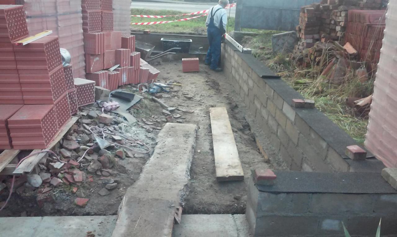 Budowa fundamentów z cegły ceramicznej, widoczny murarz w kombinezonie i poziomica, stosy bloczków i gruzu budowlanego, taśma ostrzegawcza w tle.