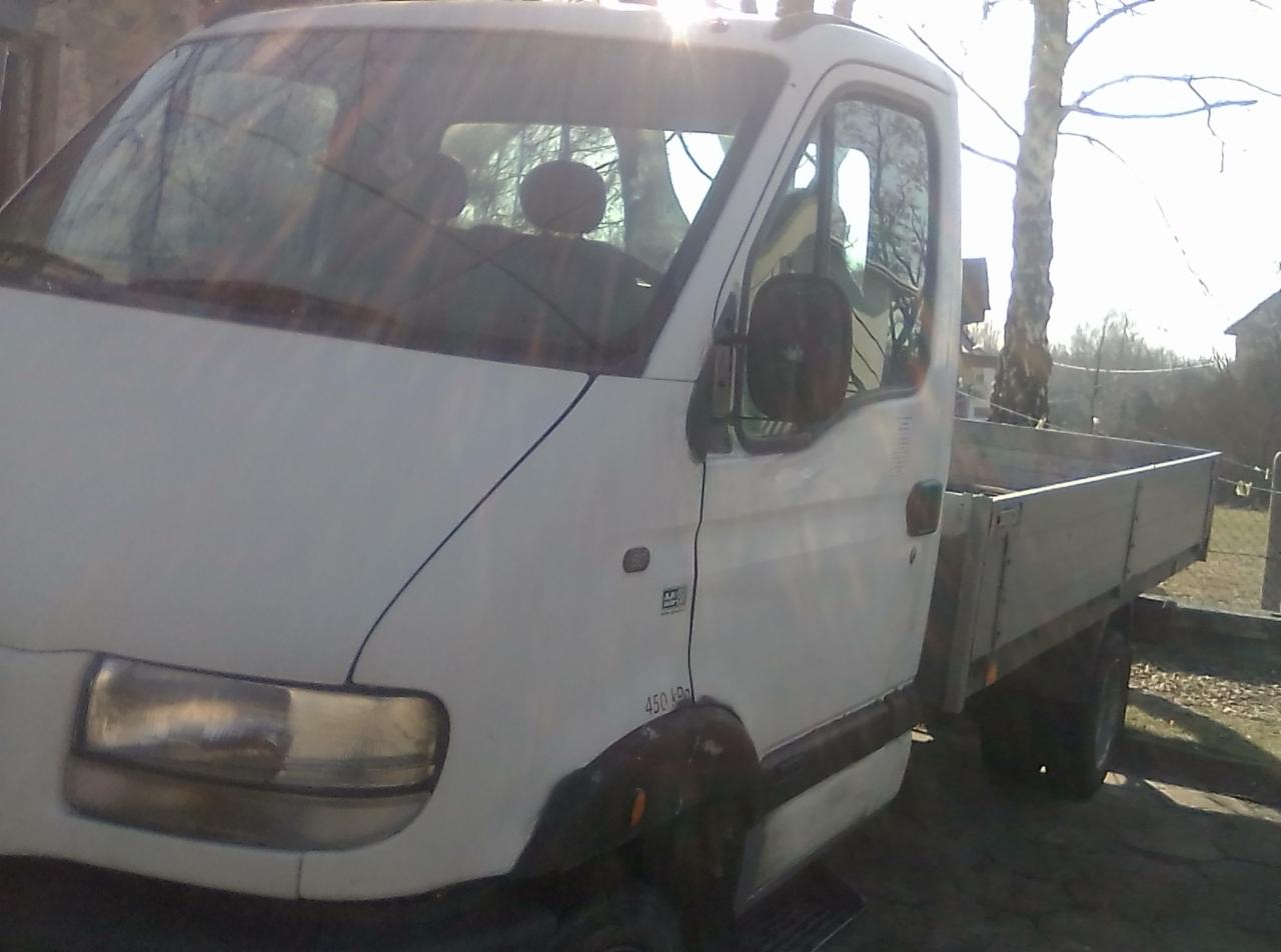 Biały samochód dostawczy z otwartą skrzynią ładunkową, zaparkowany na zewnątrz w słoneczny dzień, widoczne logo producenta na karoserii i fragment zabudowań w tle.