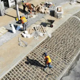 Widok z góry na robotników układających kostkę brukową na nowym chodniku, z widoczną betoniarką i narzędziami na poboczu, w słoneczny dzień.