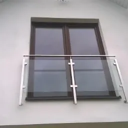 Balkon francuski z szklaną balustradą i metalowymi słupkami na tle białej elewacji domu