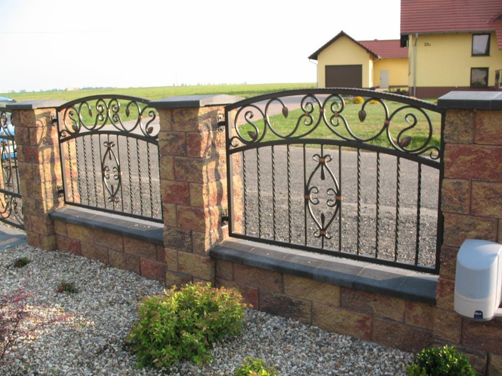 Kute ogrodzenie z dekoracyjnymi elementami roślinnymi i ceglanymi słupkami z szarymi nakrywami, widoczny automat do bramy.