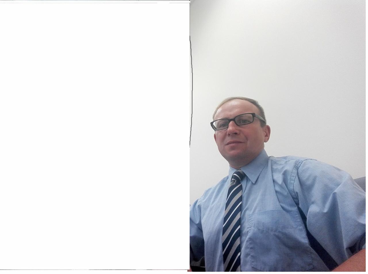 Portret mężczyzny w okularach, błękitnej koszuli i krawacie w paski na tle białej ściany, z dużą białą przestrzenią po lewej stronie kadru.