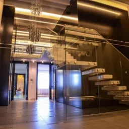 Nowoczesne schody z przezroczystego szkła z podświetlanymi stopniami, zamontowane w przestronnym holu z ciemnymi ścianami i wiszącymi kryształowymi żyrandolami.