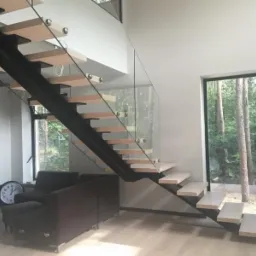 Nowoczesne schody z drewnianymi stopniami, czarną metalową konstrukcją i szklaną balustradą, widok na las przez duże okno.