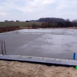 Świeżo wylana, gładka płyta betonowa fundamentowa z wystającymi prętami zbrojeniowymi, widoczne rury instalacyjne i otaczający teren budowy z ziemią.