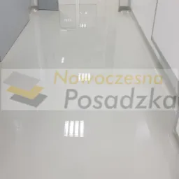 Błyszcząca, jasnoszara posadzka w korytarzu przemysłowym, odbijająca światło lamp sufitowych, z widocznymi drzwiami po bokach i w tle, oraz logo firmy naklejone na podłogę.