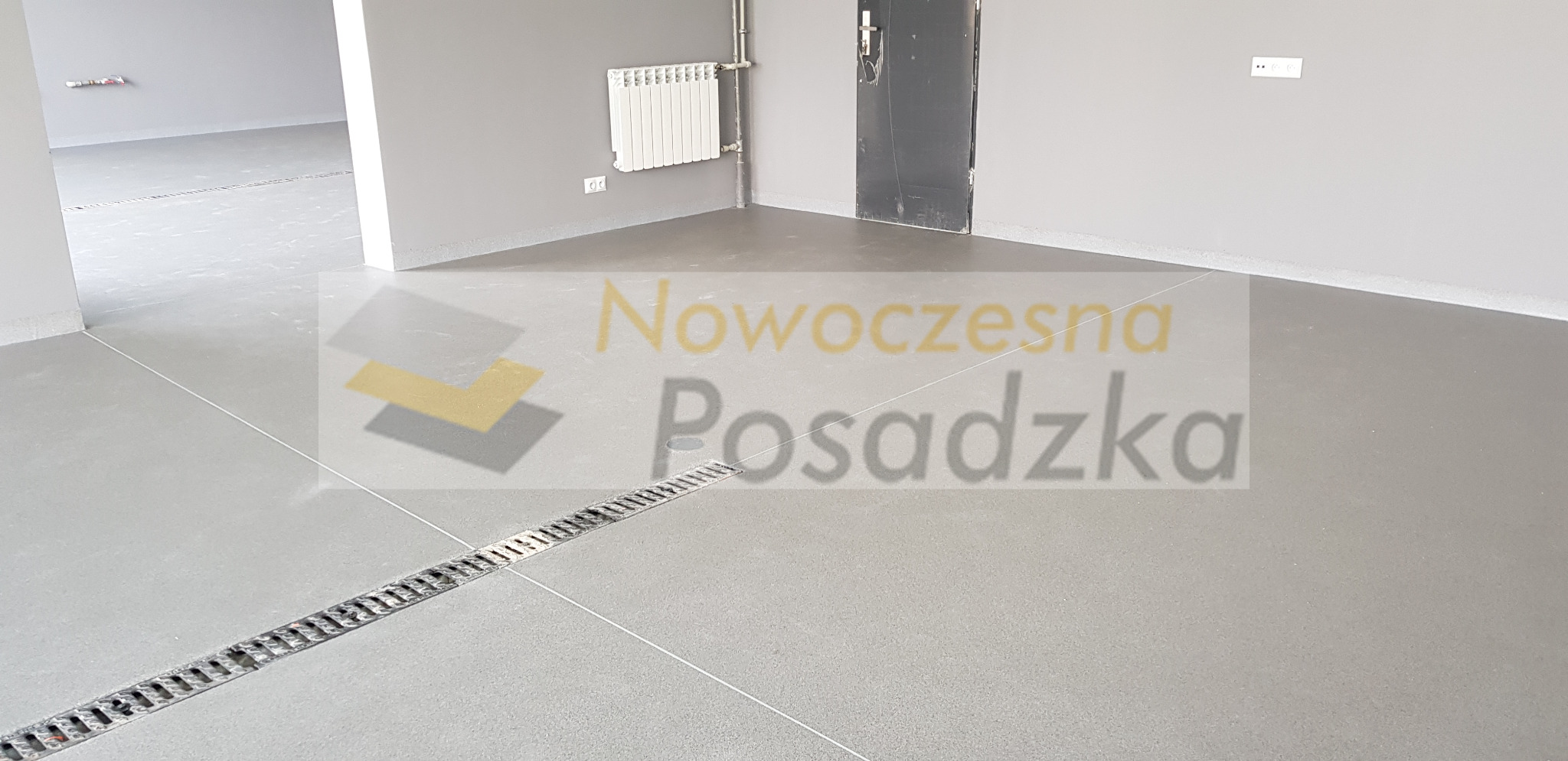 Szara, gładka posadzka z odpływem liniowym w nowym pomieszczeniu, widoczny grzejnik na ścianie i surowa, czarna futryna drzwi.