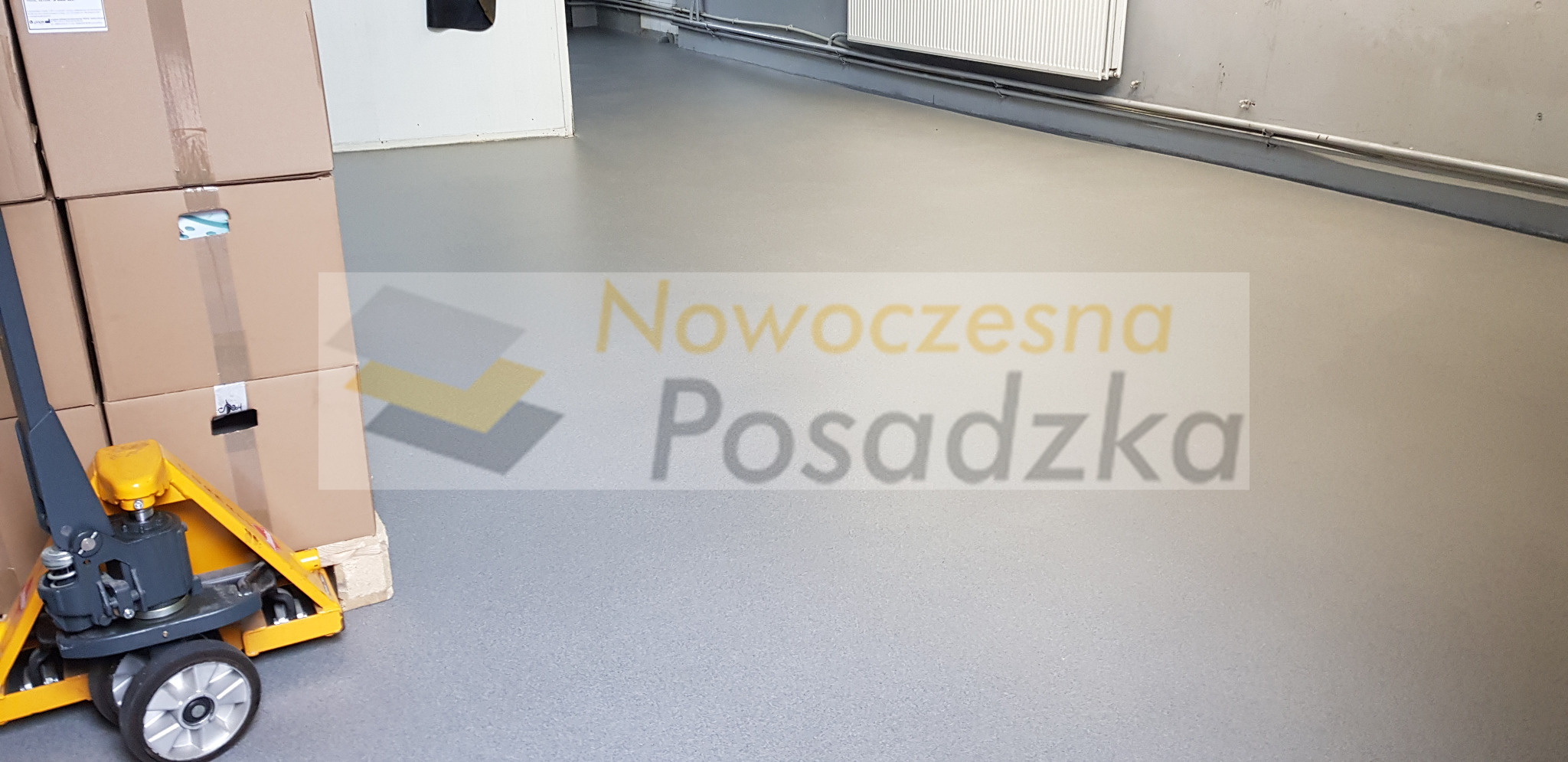 Szara, gładka posadzka w hali magazynowej z paleciakiem i stosem kartonów po lewej stronie. W tle widoczny grzejnik i rury przy ścianie.