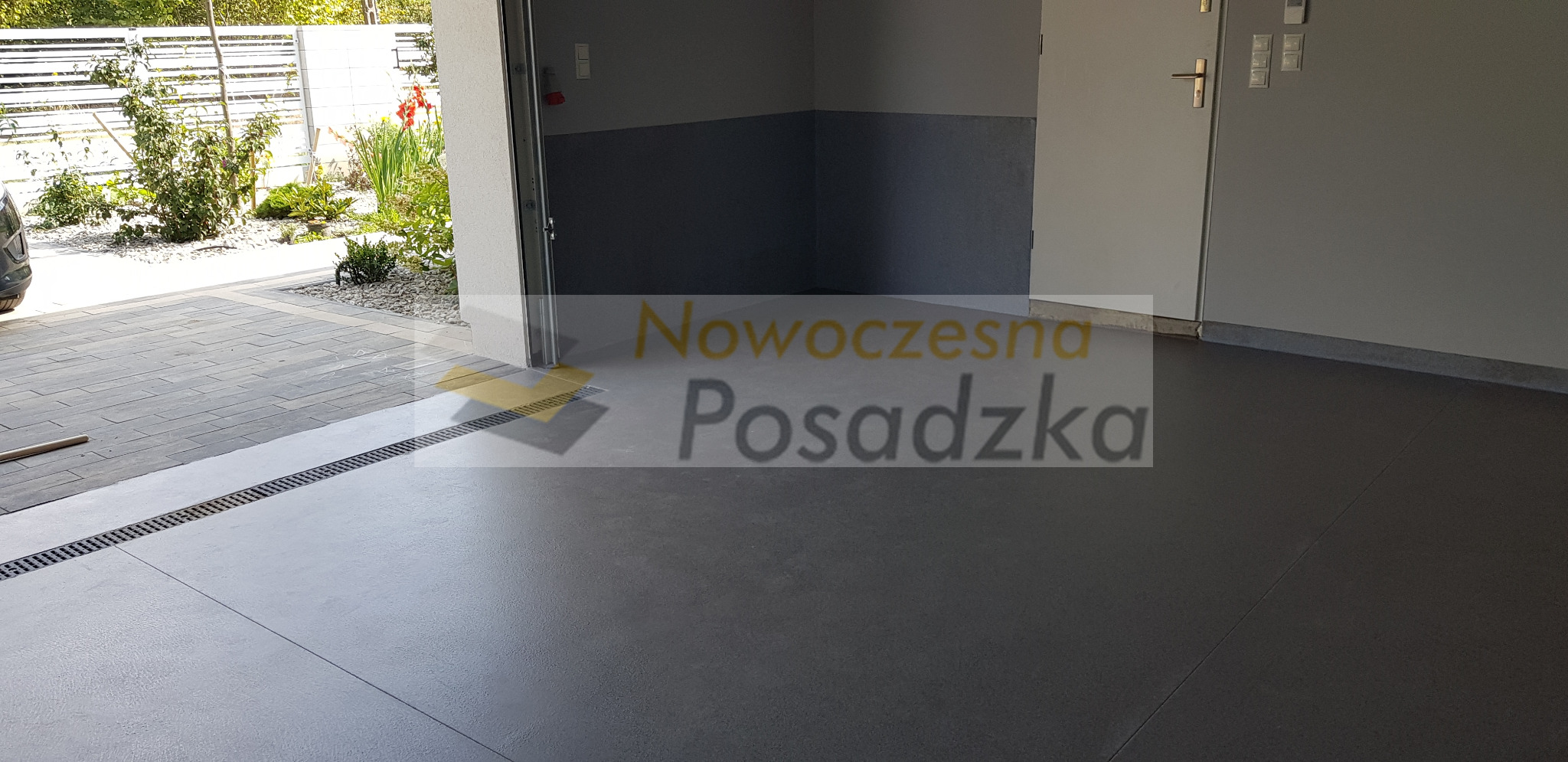 Szara, gładka posadzka w garażu, z widocznym odpływem liniowym przy wjeździe i fragmentem kostki brukowej na zewnątrz.