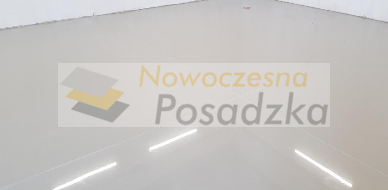 Jasnoszara, gładka posadzka z delikatnym połyskiem, widoczne refleksy świetlne, w tle rozmyte logo firmy.