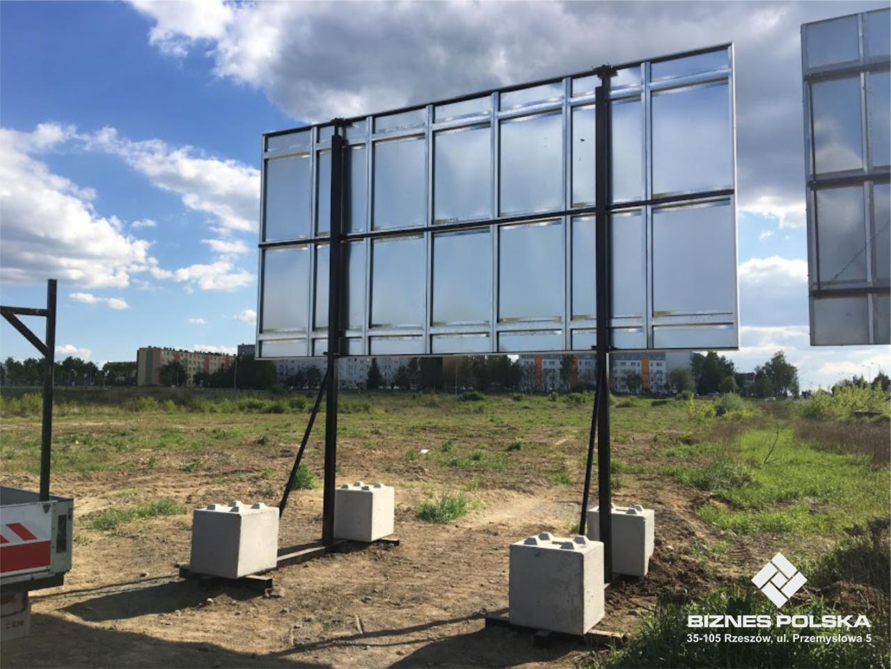 Duża tablica reklamowa o wymiarach 600x300 cm, z metalową konstrukcją wspartą na betonowych blokach, ustawiona na niezagospodarowanym terenie z widokiem na budynki mieszkalne w tle.