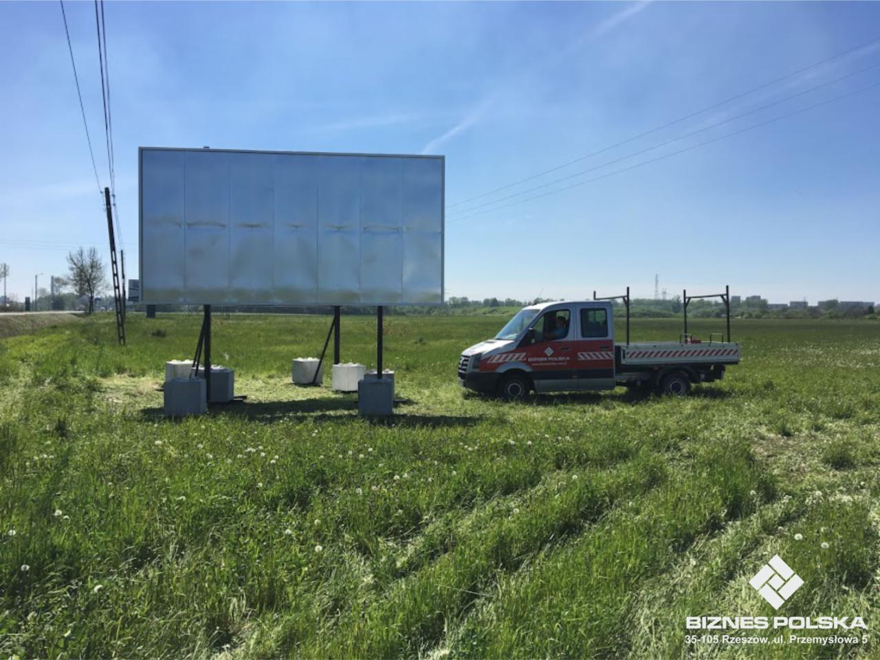 Duża, metalowa tablica reklamowa o wymiarach 600 x 300 cm, ustawiona na betonowych podstawach na łące, z widocznym samochodem dostawczym w tle.