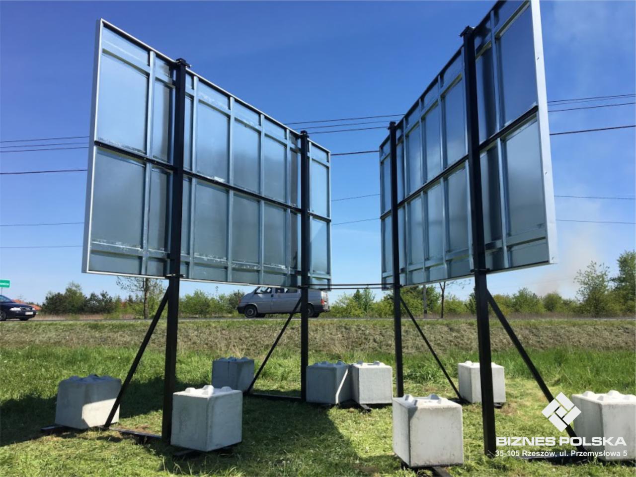 Dwie metalowe tablice przestawne o wymiarach 600x300 cm, wsparte na czarnych stalowych konstrukcjach i betonowych blokach, ustawione na trawiastym terenie przy drodze w słoneczny dzień.