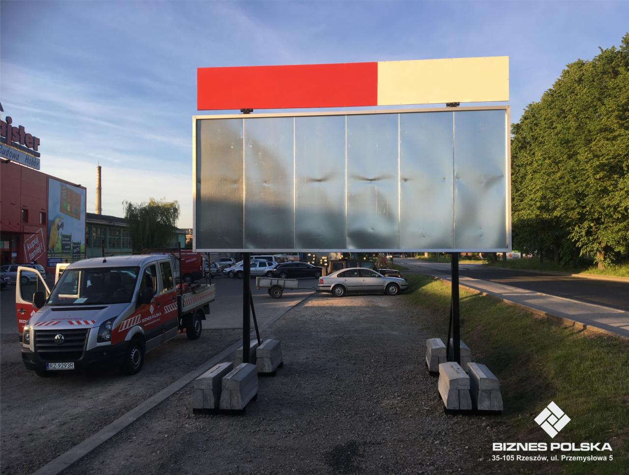 Dwustronna tablica reklamowa 502 x 240 cm z nadstawką, umieszczona na betonowych podstawach, z zaparkowanym samochodem osobowym i dostawczym w tle.