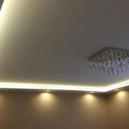 Nowoczesny sufit podwieszany z oświetleniem LED i kryształowym żyrandolem, widok z dołu, beżowe ściany i firanka w tle.