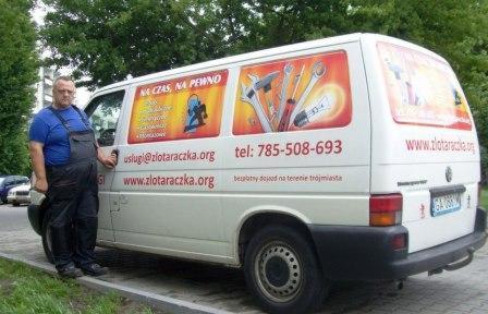 Mężczyzna w kombinezonie roboczym stoi obok białego samochodu dostawczego z reklamą usług 'złota rączka' i numerem telefonu, na tle zieleni.