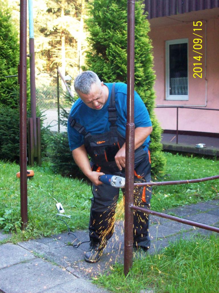 Mężczyzna w niebieskiej koszulce i roboczym kombinezonie szlifuje metalową barierkę szlifierką kątową, iskry lecą na trawnik i chodnik, w tle zielona roślinność i fragment budynku.
