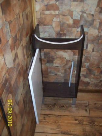 Brązowa szafka pod umywalkę z otwartymi białymi drzwiczkami, widoczna rura instalacyjna i fragment ściany z imitacją kamienia, ujęcie z niskiej perspektywy.