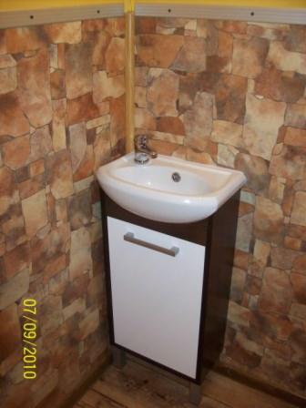 Umywalka narożna z szafką w łazience wykończonej panelami imitującymi kamień, widoczny kran i data na zdjęciu.