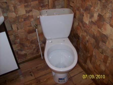 Biała, kompletna toaleta kompaktowa z widoczną rurką doprowadzającą wodę, na tle ściany wyłożonej płytkami imitującymi kamień i drewnopodobną podłogą.