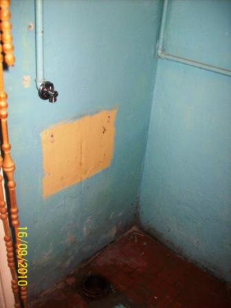 Narożnik łazienki z niebieskimi ścianami, fragmentem rury z zaworem, załatana żółtą farbą ściana, odpływ podłogowy i fragment drewnianej balustrady.