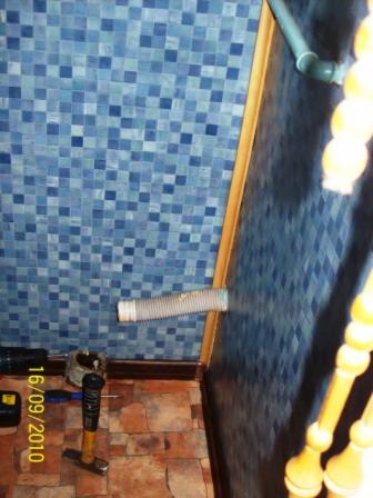 Wąż odpływowy wystający ze ściany wyłożonej niebieską mozaiką, widoczne narzędzia na podłodze z płytek oraz fragment schodów z drewnianą balustradą.