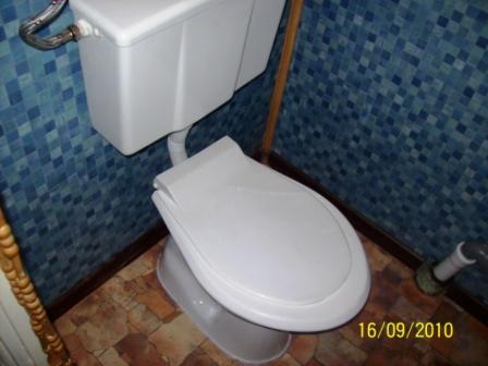 Biała toaleta kompaktowa w łazience z niebieską mozaiką na ścianach i brązową podłogą z płytek, widoczny fragment rury odpływowej.