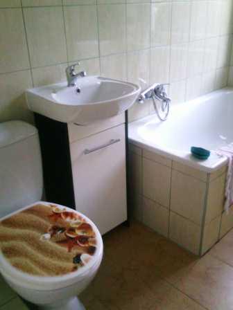 Umywalka z szafką, wanna obudowana płytkami i toaleta z deską imitującą piasek i muszle w łazience z beżowymi płytkami.